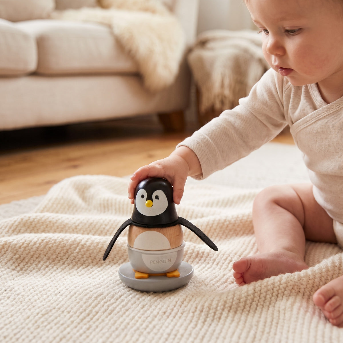 Baby sitting on a cream blanket reaching for the wooden penguin stacker