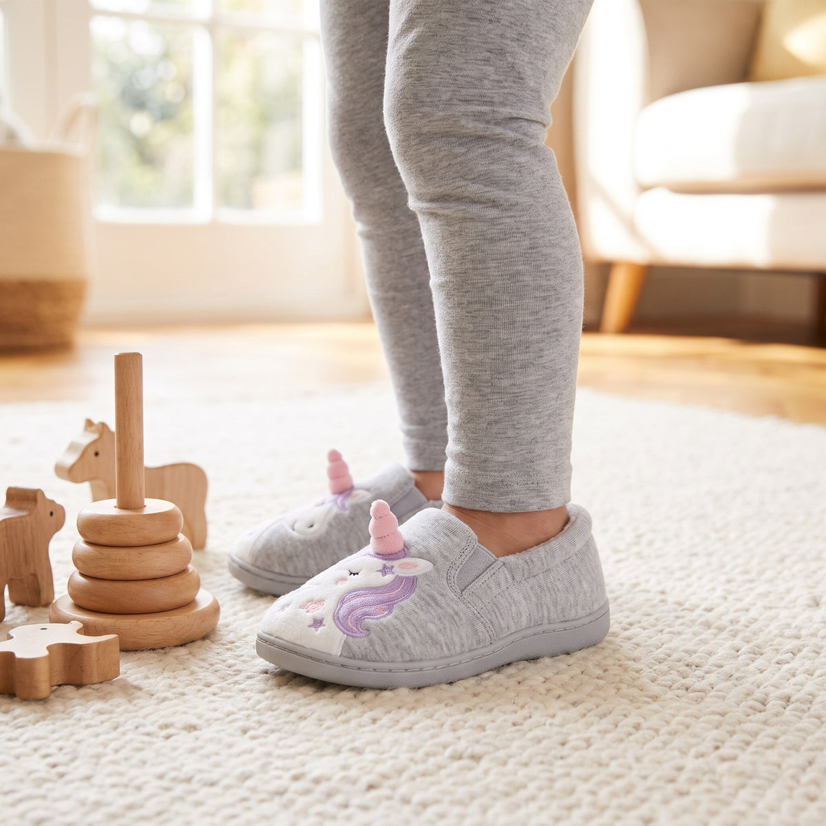 Child's feet in grey unicorn slippers on a playroom rug beside wooden toys
