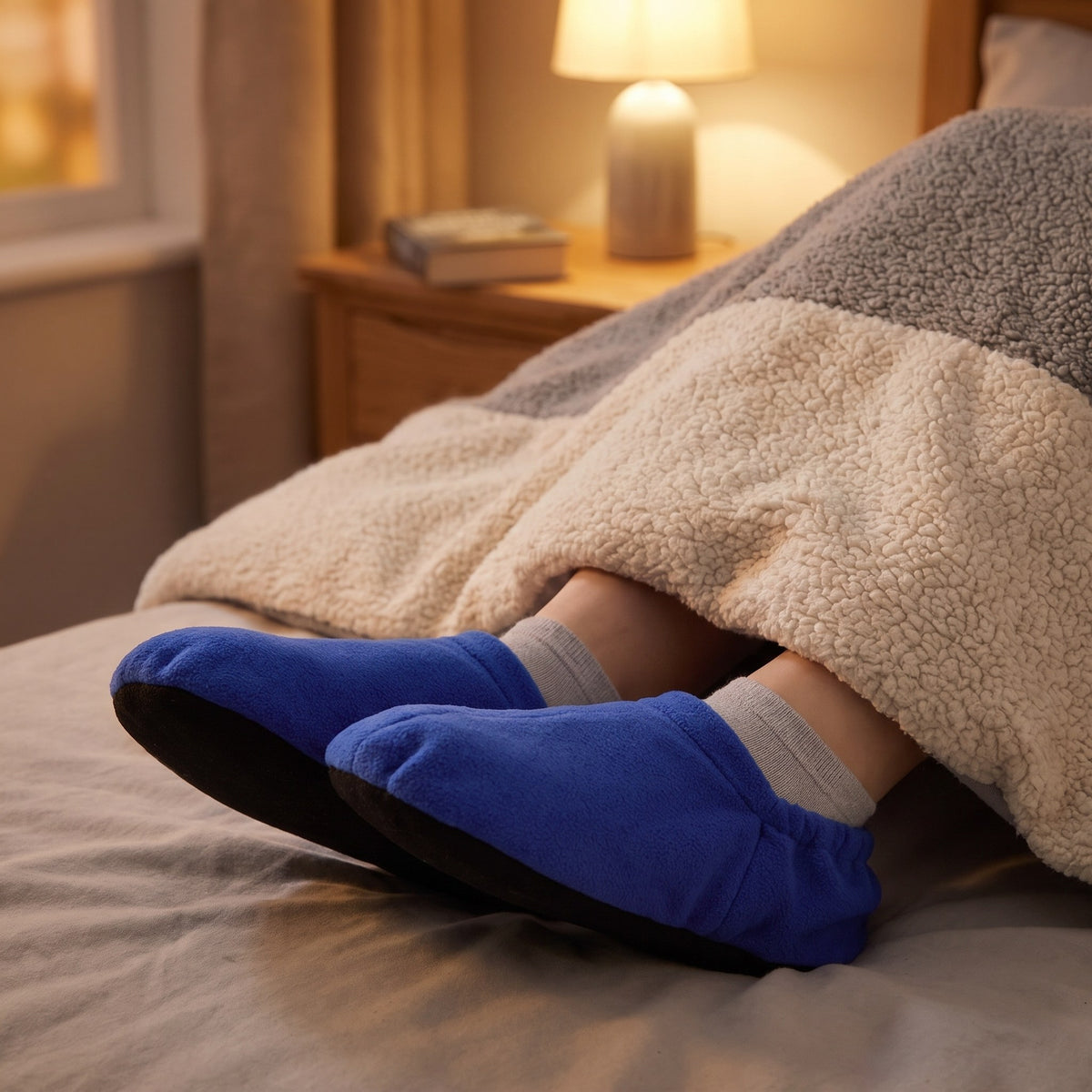 Feet in blue heated fleece foot warmers on a bed at night