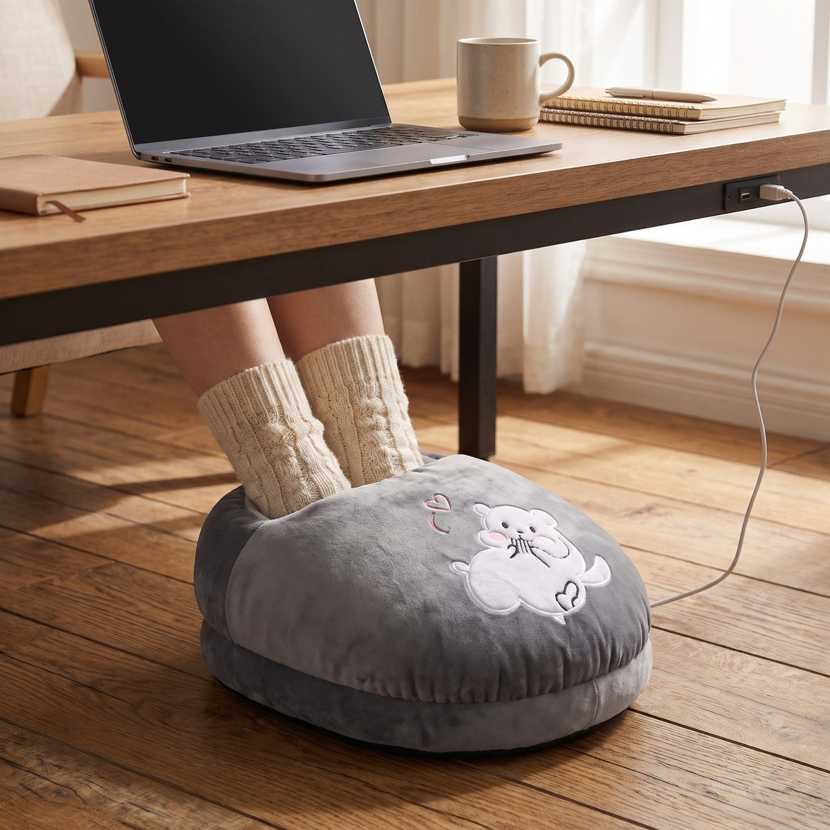 Feet in knitted socks inside a grey mouse heated foot warmer under a desk